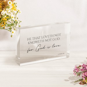 May include: A clear acrylic block with the text "HE THAT LOVETH NOT KNOWETH NOT GOD: for God is love. 1 JOHN 4:8" printed on it. The block is sitting on a white surface with flowers on either side.