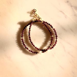 May include: A double-strand bracelet made with small, dark red garnet beads and a single white pearl charm. The bracelet has a gold clasp.