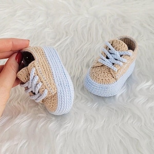 Könnte beinhalten: Ein Paar handgefertigte Babysneaker. Die Schuhe sind mit beigefarbenem Garn gehäkelt und haben hellblaue Sohlen und Schnürsenkel. Die Sneaker werden auf einer weißen, flauschigen Oberfläche präsentiert.