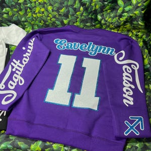 Może przedstawiać: Fioletowa bluza z białym napisem "Sagittarius" na lewym rękawie, "Evelyn Season" na prawym rękawie i "11" na plecach. Liczba 11 jest obwiedziona niebieskim brokatem.