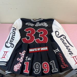 Może przedstawiać: Czarno-biała kurtka bejsbolowa z czerwonymi literami z brokatu. Kurtka ma napis "Ms. Snkr" i numer "33" na plecach. Prz&oacute;d kurtki ma napis "Gemini" na lewym rękawie i "Season Gemini" na prawym rękawie. Kurtka ma r&oacute;wnież czarną plisowaną sp&oacute;dnicę z napisem "Ms. Snkr" i "Est 1991" z przodu. Sp&oacute;dnica ma r&oacute;wnież czerwoną etykietę z napisem "Crafts by Snkr".