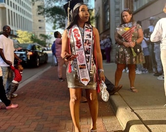 May include: A woman in a silver dress and graduation cap and gown poses for a photo with a personalized graduation sash that says "Ashley" and has photos on it. The sash is red, white, and gold.