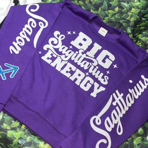 Może przedstawiać: Fioletowa bluza z białymi błyszczącymi literami, na kt&oacute;rej z przodu napisano "Big Sagittarius Energy", a na rękawie "Sagittarius".