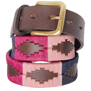 May include: A brown leather belt with a gold buckle and a woven pink, blue, and white strap. The strap has a brown leather patch with a polo player design.