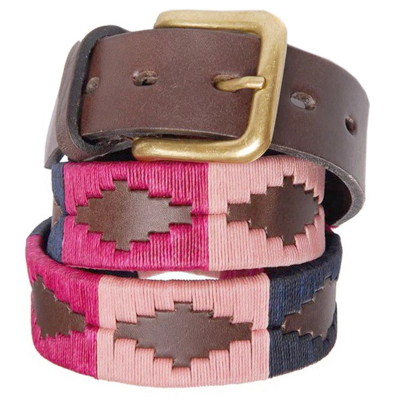 May include: A brown leather belt with a gold buckle and a woven pink, blue, and white strap. The strap has a brown leather patch with a polo player design.