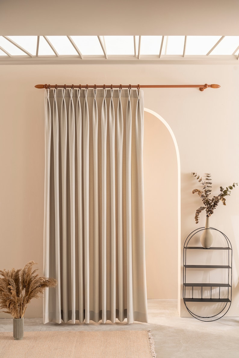 May include: A light beige curtain with pleats hangs from a wooden rod in a room with a skylight. The curtain is in front of an arched doorway with a black metal shelf unit to the right. The shelf unit has three shelves and a vase with dried branches on the top shelf.