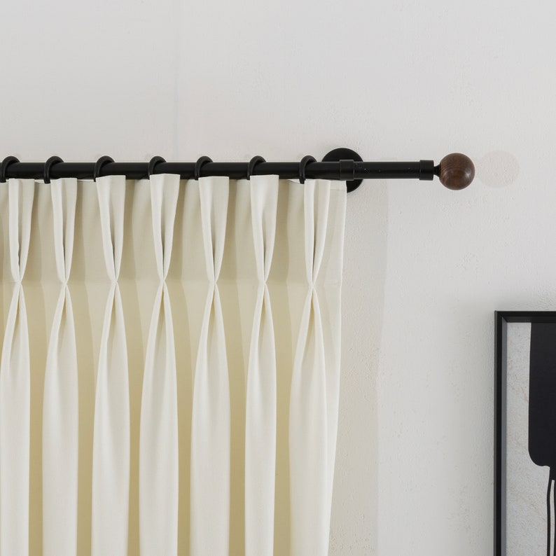 May include: White curtains with pleats hanging from a black metal curtain rod with wooden finials.
