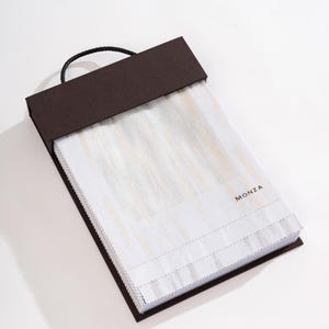 May include: A brown fabric swatch book with a black handle. The book is filled with white fabric swatches with a subtle striped pattern. The word "MONZA" is printed in gold on the front of the book.