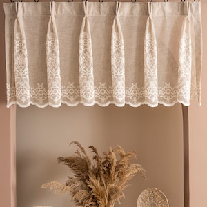 May include: A beige valance with white lace trim hangs from a black curtain rod. The valance is decorated with a white floral pattern. Below the valance, a wooden table displays a vase with dried pampas grass and two decorative sculptures.