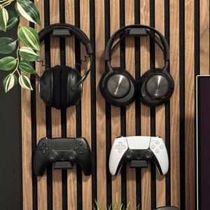 Puede incluir: Una pared de madera negra y marrón con un estante que sostiene un teclado negro. Hay dos pares de auriculares negros colgados de ganchos, dos mandos de videojuegos negros, un router negro, una planta verde en maceta blanca, un altavoz negro y una base de carga inalámbrica negra y blanca sobre una mesa de madera.