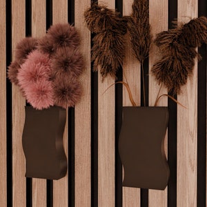 May include: Three black vases with different dried flowers hanging on a wood slat wall. The flowers are pink, brown, and white.