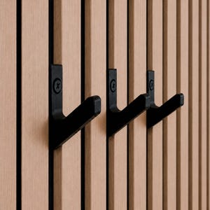 Könnte beinhalten: Drei mattschwarze Metallhaken, die an einem Wandpaneelsystem aus vertikalen, hellbraunen Holzlatten befestigt sind. Jeder Haken ist mit einer sichtbaren schwarzen Schraube befestigt und hat ein einfaches L-förmiges Design.