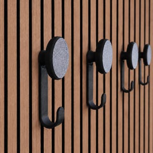 Può includere: Una serie di quattro ganci a parete con base in metallo nero a forma di J e faccia frontale circolare con finitura grigia testurizzata simile alla pietra, fissati a una parete a pannelli con listelli verticali in legno marrone chiaro.