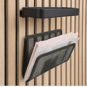 Op de afbeelding: Een zwart metalen tijdschriftenrek en plank bevestigd aan een licht houten wandpaneel. Het rek heeft een gaasontwerp en bevat kranten. De plank heeft een eenvoudige, rechthoekige vorm. Het ontwerp is modern en minimalistisch.