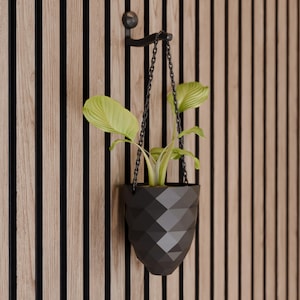 Può includere: Un vaso da fiori sospeso geometrico nero con una pianta verde appesa a una catena nera su una parete a doghe di legno.