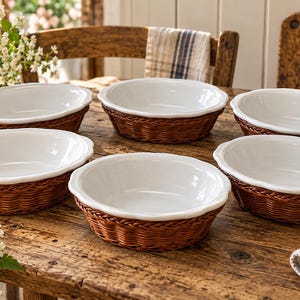 May include: A set of five round white ceramic baking dishes, each nestled within a woven brown wicker basket base. The dishes feature smooth, glossy white interiors and scalloped edges, providing a contrast to the textured, natural brown wicker.