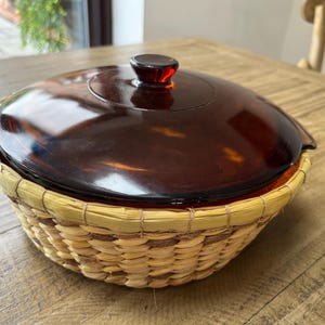 May include: A round amber-colored glass casserole dish with a matching lid, set inside a woven light-tan straw basket holder. The glass lid features a central knob handle, and the basket has a reinforced rim with a natural, textured weave pattern.