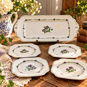 May include: A rectangular serving platter and four matching square plates, all crafted from white ceramic. Each piece features a hand-painted design of purple flowers with green leaves and a delicate green vine border along the scalloped edges.