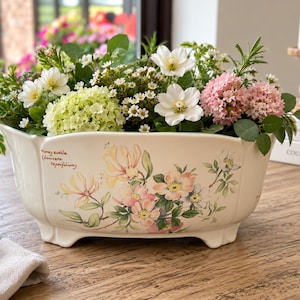 May include: An off-white ceramic planter with a scalloped rim and four small feet. The exterior features a botanical illustration of honeysuckle flowers with the text "Honeysuckle (Lonicera caprifolium)" printed in brown script on the side.