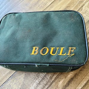 May include: A rectangular dark green zipped storage case with rounded corners and black piping along the edges. The word BOULE is embroidered in yellow capital letters on the front surface of the fabric case, which has a textured weave.