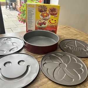 May include: A Sun Select Special fantasy bakeware set featuring a red circular springform pan base and four interchangeable metal inserts with embossed designs of a teddy bear, a car, a smiley face, and a flower, accompanied by the original box.