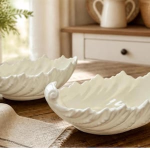 May include: Two white ceramic bowls with a decorative leaf-shaped design. Each bowl features a textured, scalloped edge that mimics the appearance of foliage, with a curled detail at one end. The bowls have a glossy finish and an oval shape.