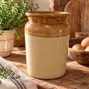 May include: A cylindrical stoneware crock featuring a two-tone design with a glossy brown glazed rim and upper section, transitioning into a smooth, matte cream-colored base. The vessel has a thick, rounded top edge and a sturdy, straight-sided body.