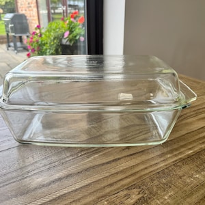 Puede incluir: Una fuente de horno rectangular de vidrio transparente con su tapa de vidrio a juego. El recipiente cuenta con asas laterales integradas y una superficie lisa y transparente, diseñada para su uso en el horno. La tapa encaja sobre la base.