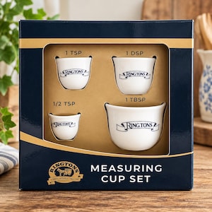 May include: A boxed set of four white ceramic measuring cups with blue text. The cups are labeled 1 TSP, 1 DSP, 1/2 TSP, and 1 TBSP, each featuring the Ringtons logo. The dark blue box displays the text Ringtons Measuring Cup Set.