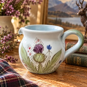 以下が含まれることがあります： 緑、紫、青の色合いでアザミの花が手描きされた、水色の陶器製クリーマーピッチャー。丸みを帯びた本体に小さな注ぎ口があり、上端に淡い緑色の色合いが施された曲線的なハンドルが付いています。