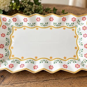 Puede incluir: Una bandeja de servir rectangular blanca con un borde ondulado y festoneado. El borde presenta un patrón repetitivo de motivos florales rojos y ramitas verdes, delimitado por una línea dorada con pequeñas estrellas amarillas en el centro.