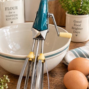 May include: A vintage manual hand mixer featuring a teal enamelled metal body, chrome-plated beaters, and cream-coloured plastic handles. The kitchen tool has a top handle for gripping and a side crank handle for rotating the two wire beaters.
