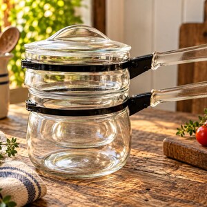 May include: A clear glass double boiler set consisting of two stackable glass pots with a matching glass lid. Each pot features a black metal band around the rim and a clear glass handle attached with black metal hardware for secure handling.