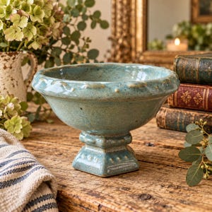 May include: A teal ceramic pedestal bowl with a speckled finish and a wide, flared rim. The bowl features a square base and decorative raised dot detailing along the rim and the pedestal. The surface has a glossy, mottled blue-green glaze.