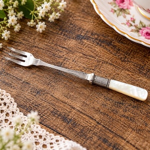 May include: A silver-toned metal dessert fork featuring three tines and a handle accented with a ribbed metal band and a smooth, white, iridescent inlay. The fork has a slender neck and a polished finish, showcasing a classic design.