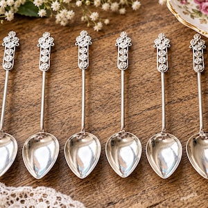 May include: A set of six silver-toned metal spoons featuring a long, slender handle topped with a decorative, vertically aligned floral pattern consisting of four circular rose-like motifs. Each spoon has a smooth, polished, teardrop-shaped bowl.