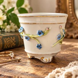 May include: A white ceramic planter with three small feet, featuring delicate raised blue floral appliques on green stems and gold trim along the rim and base. The vessel has a smooth, glossy finish and a tapered shape, showcasing intricate craftsmanship.