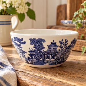 May include: A white ceramic bowl featuring a blue willow pattern that depicts traditional pagodas, willow trees, and landscape elements. The bowl has a smooth, rounded rim and a deep, circular basin, showcasing a classic transferware design.