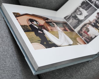 May include: A wedding album with a white cover and blue edges. The album is open to a page with a photo of a bride and groom. The bride is wearing a white dress and the groom is wearing a black suit. The photo is taken outdoors in a garden setting.