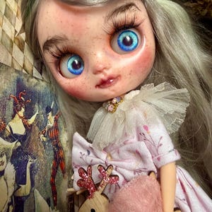 Può includere: Una bambola Blythe con grandi occhi blu, lunghe ciglia e lentiggini. La bambola ha i capelli grigi e indossa un vestito rosa con un colletto bianco a balze. Una renna di legno con corna glitterate rosse e un accessorio rosa e soffice è tenuta dalla bambola.