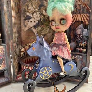 Könnte beinhalten: Eine gruselige Puppe mit mintgrünen Haaren und einem rosa Kleid sitzt auf einem blauen Schaukelpferd. Das Pferd hat goldene Stern- und Mondakzente. Der Hintergrund zeigt Zirkus-Kunstwerke und einen Clown.
