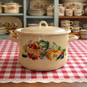 Könnte beinhalten: Cremefarbener Emaille-Kochtopf mit Deckel, mit einem farbenfrohen Obst- und Gemüsedesign. Der Topf hat zwei seitliche Griffe und einen oberen Griff. Der Topf steht auf einer rot-weiß karierten Tischdecke. Küchenutensilien.