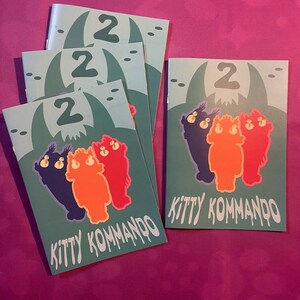 K&ouml;nnte beinhalten: Mehrere t&uuml;rkisfarbene Brosch&uuml;ren mit dem Text "KITTY KOMMANDO" und der Zahl "2". Jede Brosch&uuml;re zeigt eine Cartoon-Illustration von drei katzenartigen Figuren in Lila, Orange und Rot. Der Hintergrund zeigt eine geh&ouml;rnte Kreatur.