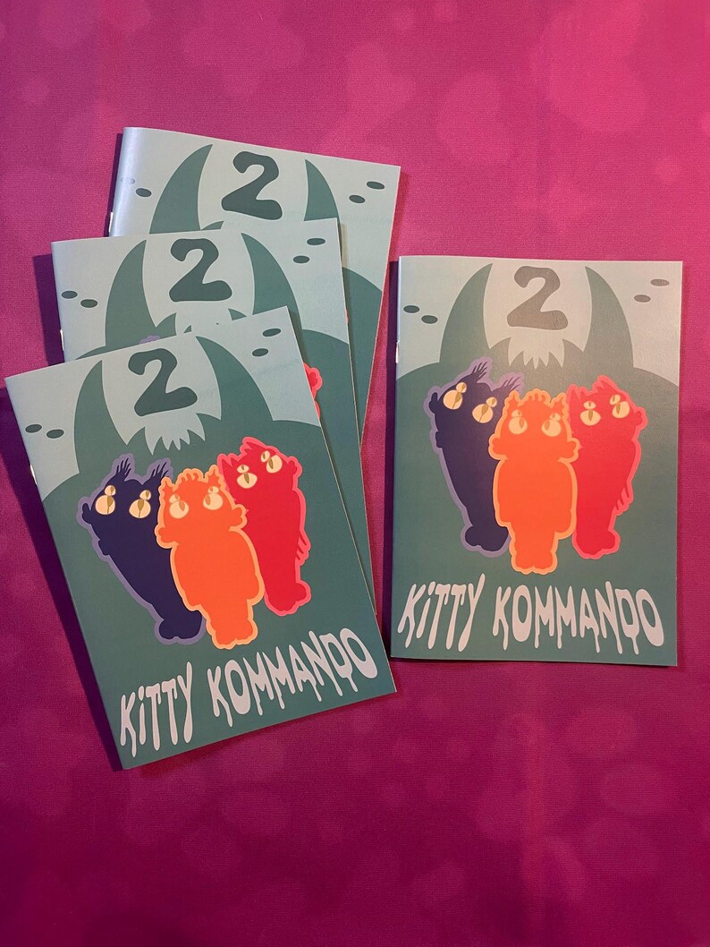 K&ouml;nnte beinhalten: Mehrere t&uuml;rkisfarbene Brosch&uuml;ren mit dem Text "KITTY KOMMANDO" und der Zahl "2". Jede Brosch&uuml;re zeigt eine Cartoon-Illustration von drei katzenartigen Figuren in Lila, Orange und Rot. Der Hintergrund zeigt eine geh&ouml;rnte Kreatur.