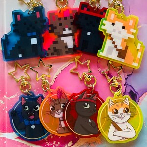 May include: Six colorful keychains featuring pixelated and cartoon cats. The keychains are attached to gold star-shaped charms. The keychains are on a pink and white marble surface.