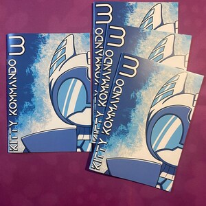 K&ouml;nnte beinhalten: Mehrere blaue und wei&szlig;e Kitty Kommando Comics liegen auf einer lila Oberfl&auml;che. Das Comic-Cover zeigt einen stilisierten Robotercharakter mit blau-wei&szlig;em Farbschema. Der Titel "Kitty Kommando" ist vertikal auf der linken Seite gedruckt.