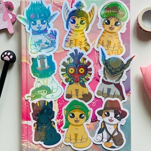 Może przedstawiać: Zestaw dziesięciu holograficznych naklejek przedstawiających różne postacie kotów w różnych kapeluszach i strojach. Naklejki znajdują się na różowo-białym tle.