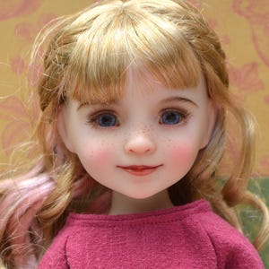 Könnte beinhalten: Eine Puppe mit blauen Augen, Sommersprossen und blondem Haar mit rosa Highlights. Die Puppe trägt ein magentafarbenes Hemd mit einem Minnie-Maus-Design in Lila und Orange. Das Gesicht der Puppe ist detailliert mit rosigen Wangen und roten Lippen.