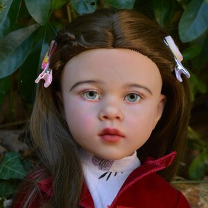 May include: A realistic doll with brown hair styled with two light pink hair clips. The doll has blue eyes, rosy cheeks, and pink lips. It wears a white high-neck shirt with a small purple floral detail and a deep red velvet jacket with buttons.