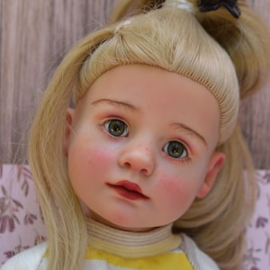 May include: A doll with blonde hair styled in a high ponytail with a black clip, featuring green eyes, rosy cheeks, and pink lips. The doll wears a yellow and white horizontal striped shirt with a round neckline and short sleeves.